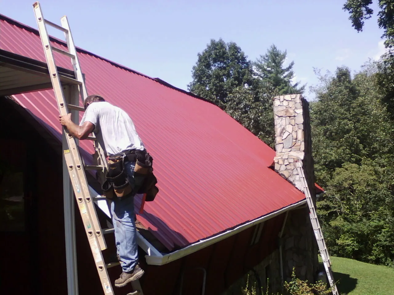 Roofer on a ladder beside a red metal roof and stone chimney — Soffit & Fascia Repair by Roofing Brothers in Irvona